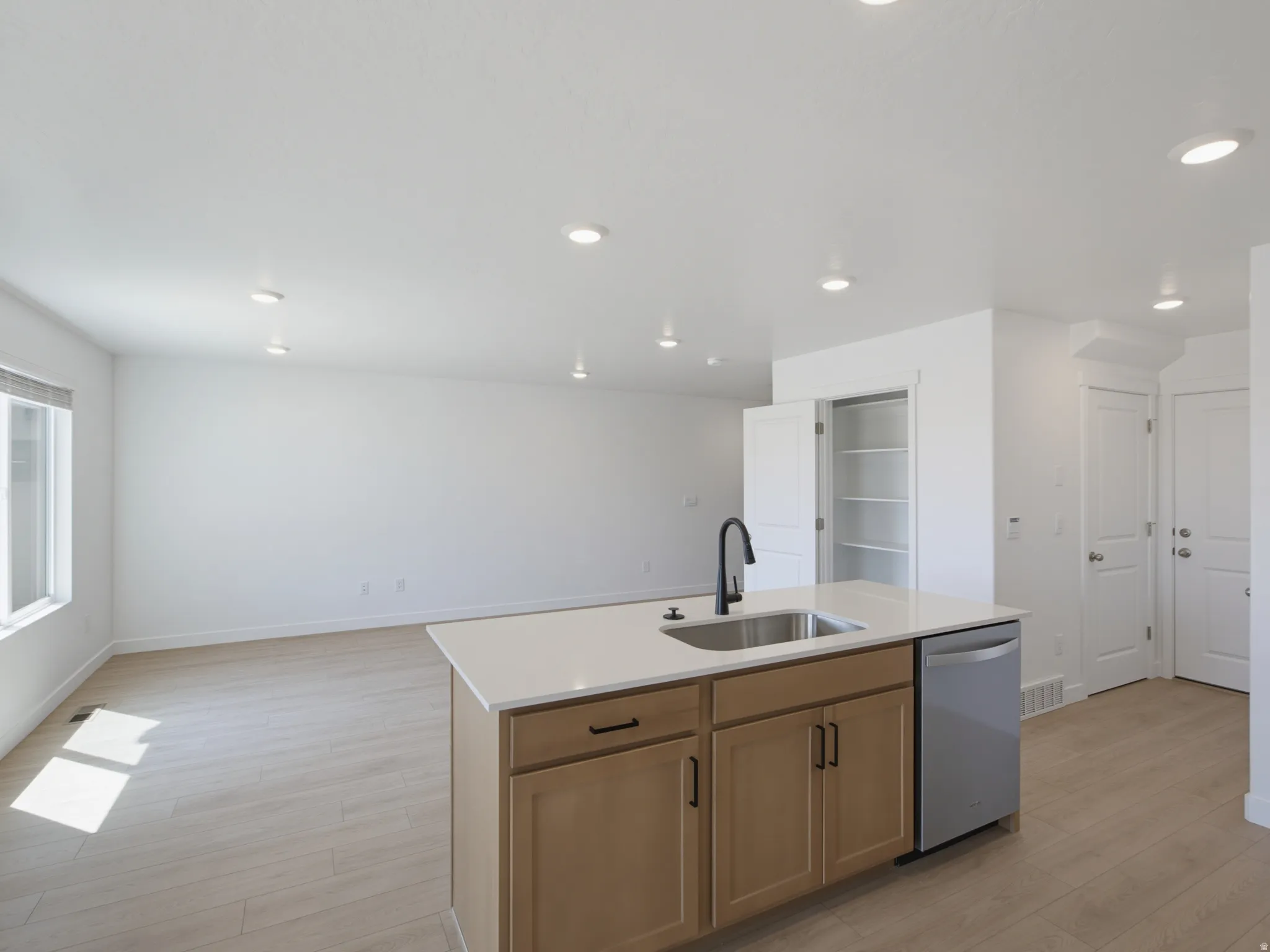 Kitchen island with integrated stainless steel sink and matte black faucet, light wood-finish cabinetry, light-toned countertops, stainless steel dishwasher, and recessed lighting
