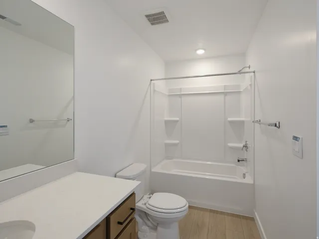 Recessed lighting illuminating a white bathtub with a shower surround, wood-finish flooring, a white toilet, and a vanity with a light-toned countertop and wood-finish cabinetry
