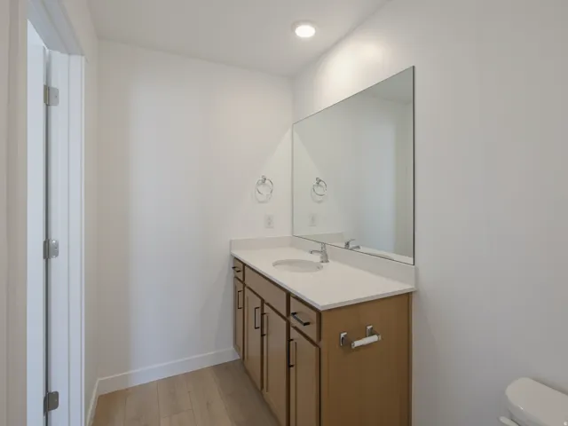 Bathroom featuring a light wood-finish vanity with a light countertop, an integrated sink, a wall-mounted mirror, and wood-finish flooring