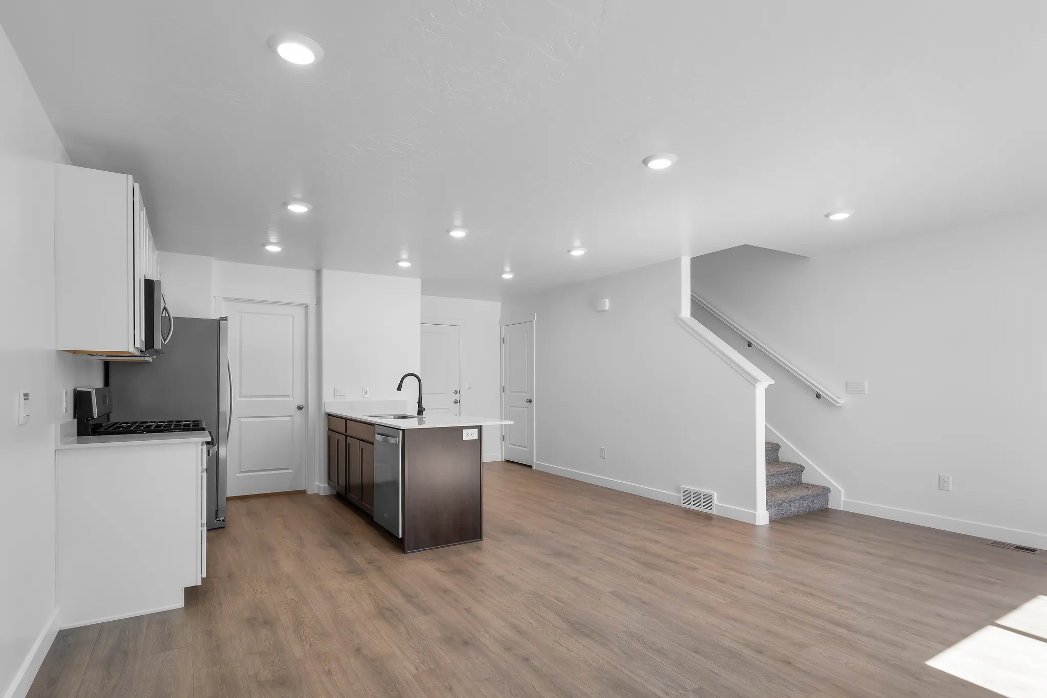 Open-concept living area featuring wood-finish flooring, recessed lighting, a kitchen island with a white countertop and dark cabinetry, stainless steel appliances, and a carpeted staircase with a white railing
