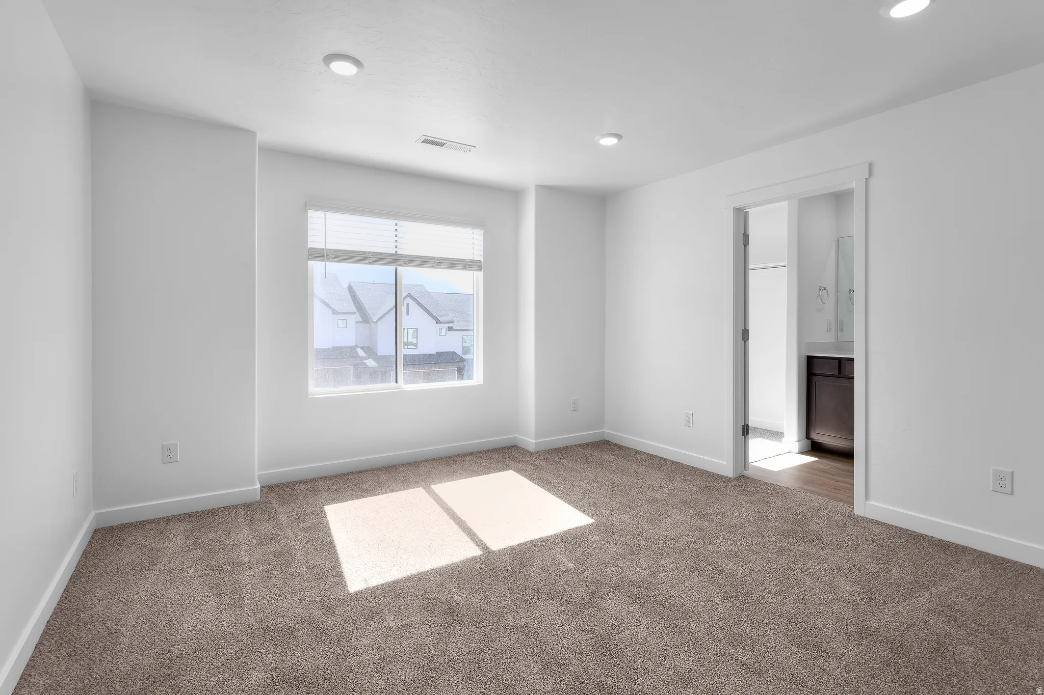 Recessed lighting, neutral carpeting, window with blinds, white walls, and a doorway opening to a bathroom with a dark wood-finish vanity