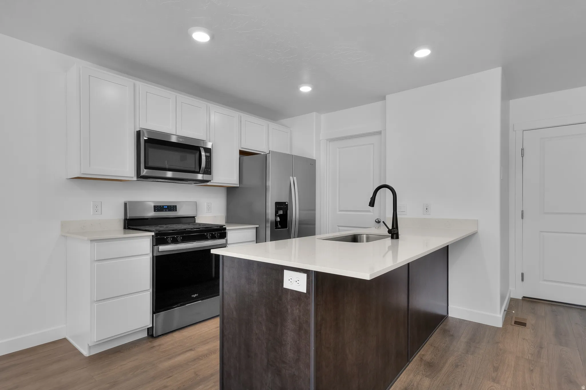 Kitchen featuring white cabinetry, stainless steel appliances, a center island with a dark wood-finish base, light-toned countertops, and wood-finish flooring