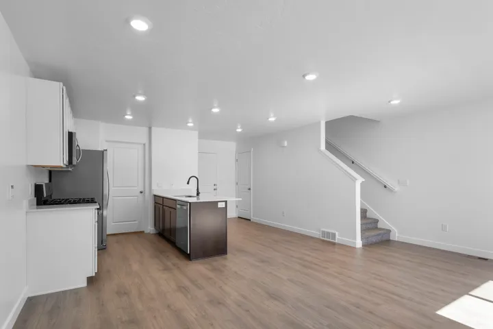 Open-concept living area featuring wood-finish flooring, recessed lighting, a kitchen island with a white countertop and dark cabinetry, stainless steel appliances, and a carpeted staircase with a white railing