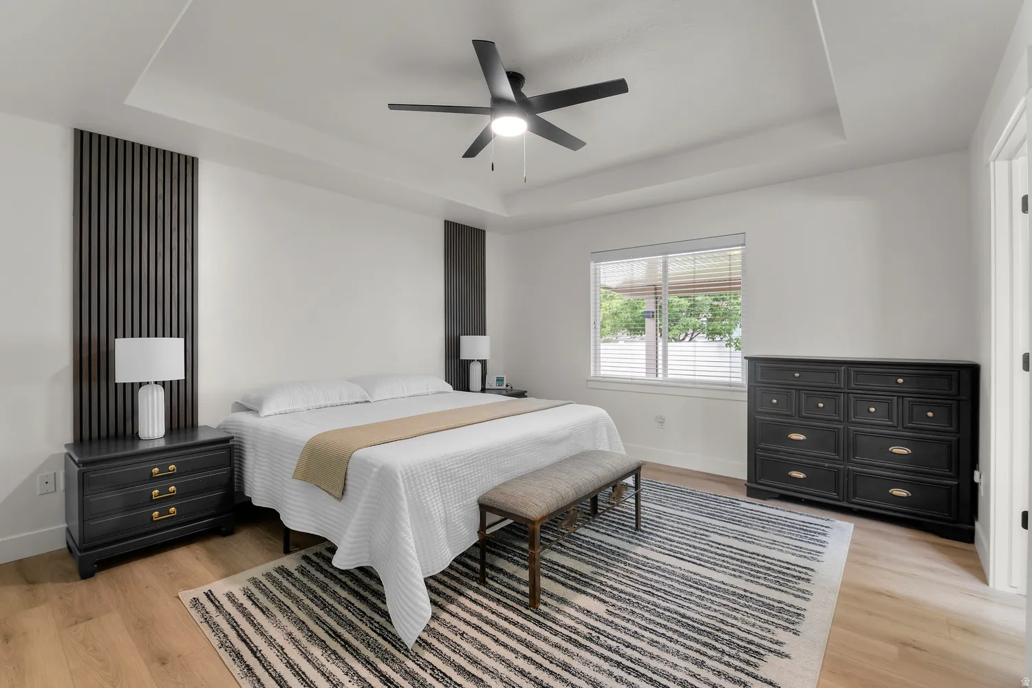 Master bedroom with Recessed tray ceiling with a ceiling fan, wood-finish flooring, and a large window with blinds