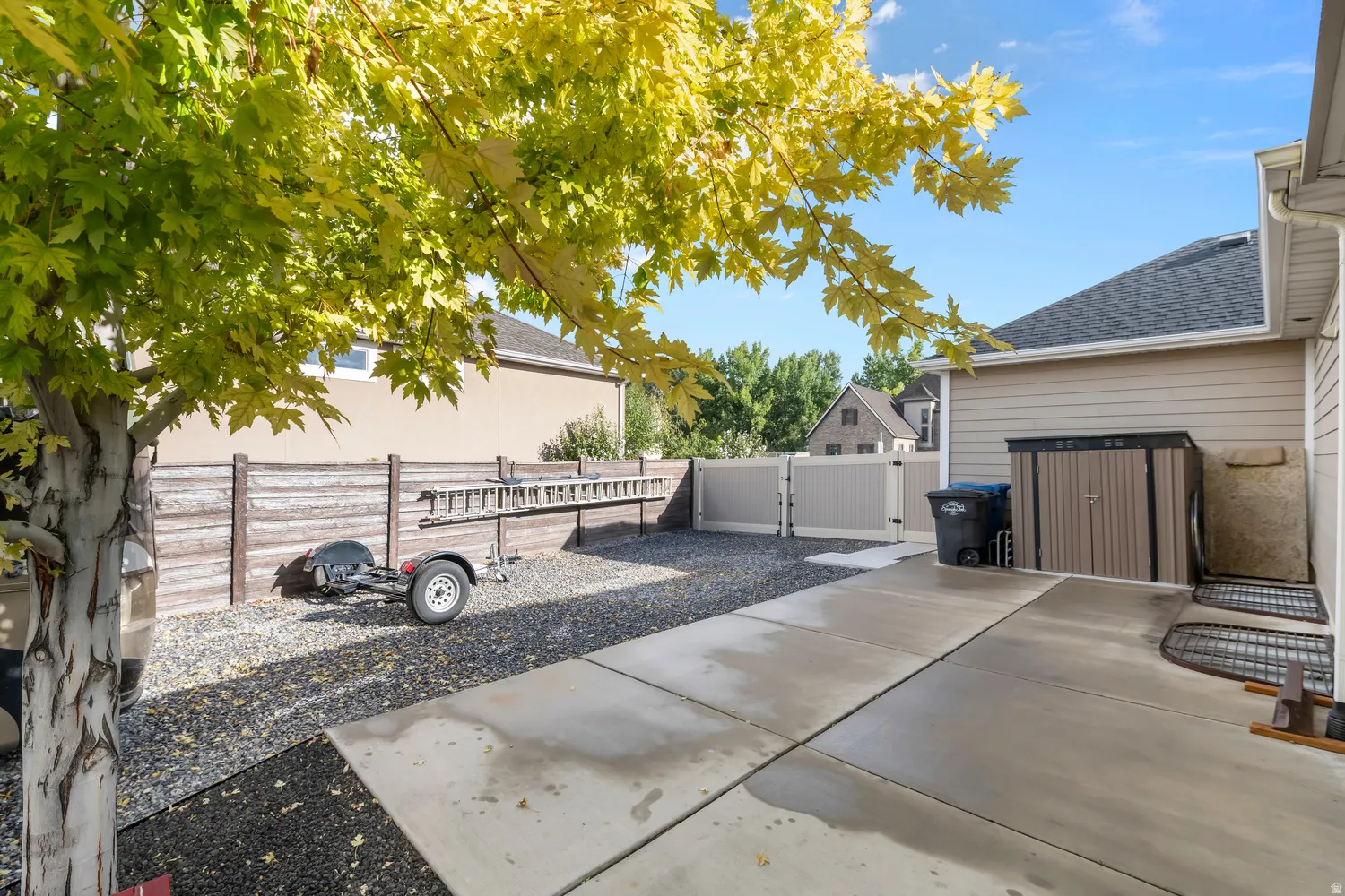 Concrete patio leading to a gravel area with a privacy fence and a white vinyl gate. 2 sheds
