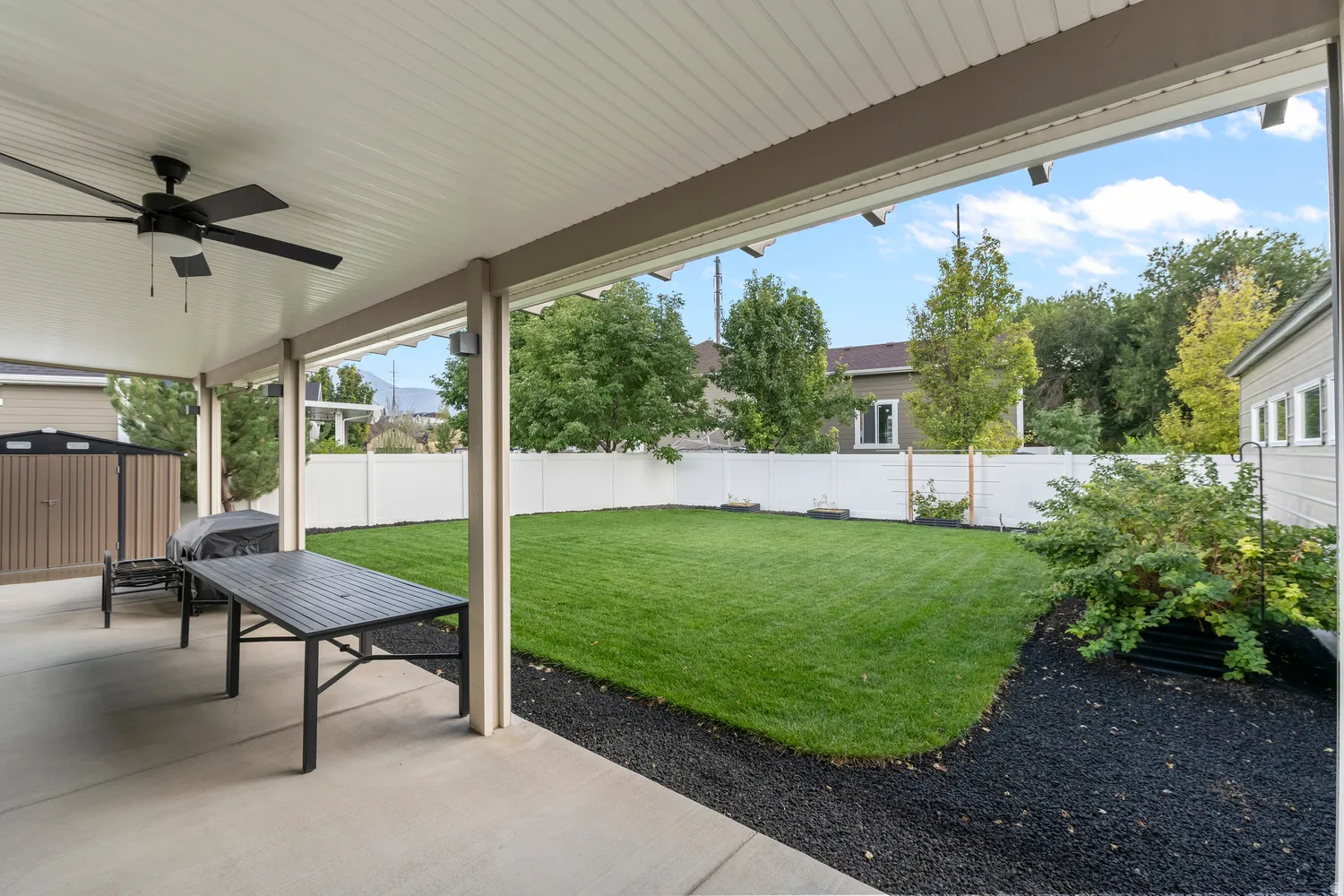 Covered concrete patio with ceiling fan, expansive fenced lawn, and perimeter landscaping