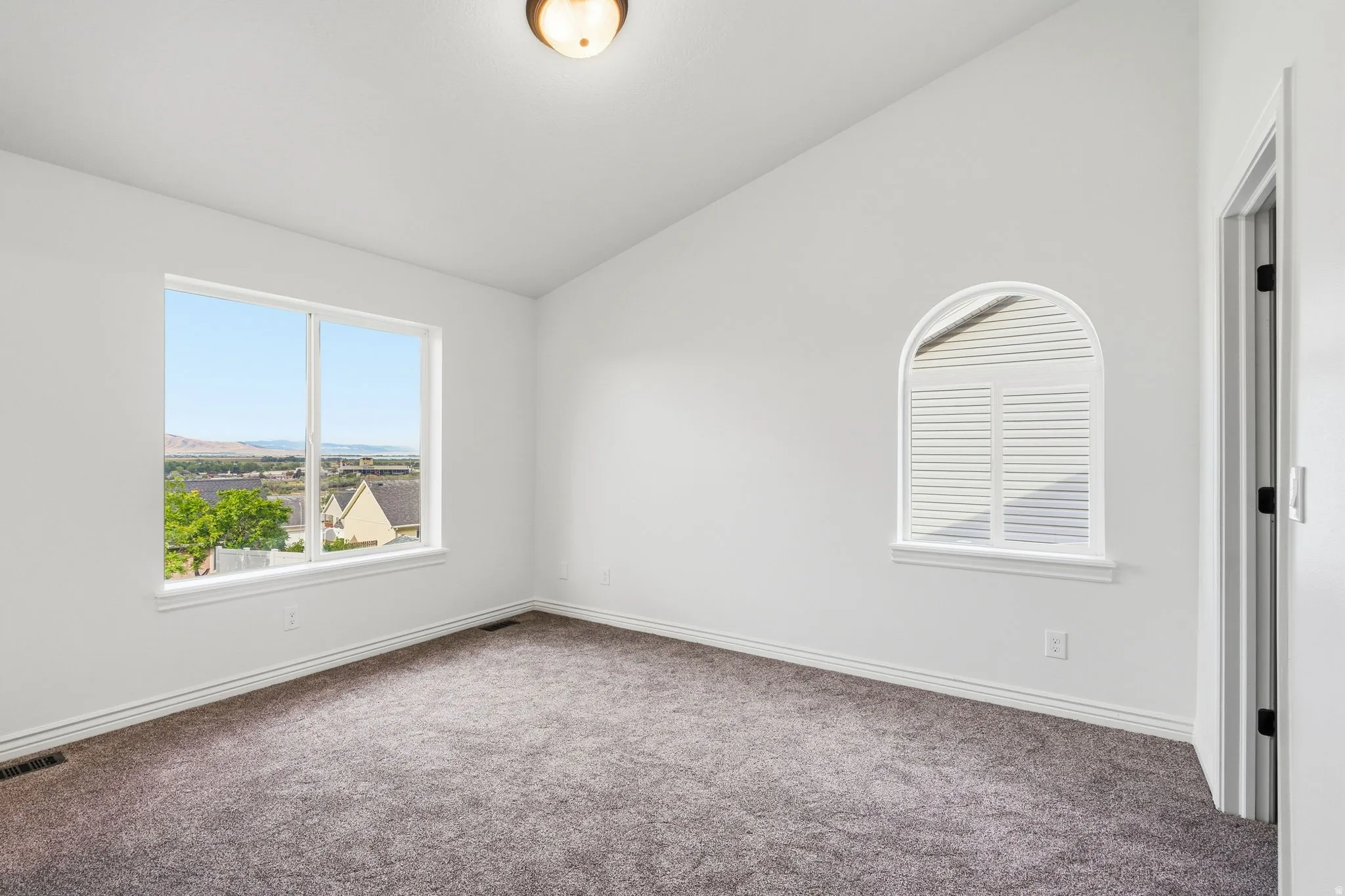 Vaulted ceiling, two windows, arched window with plantation shutters, wall-to-wall carpeting, and a flush-mount ceiling light