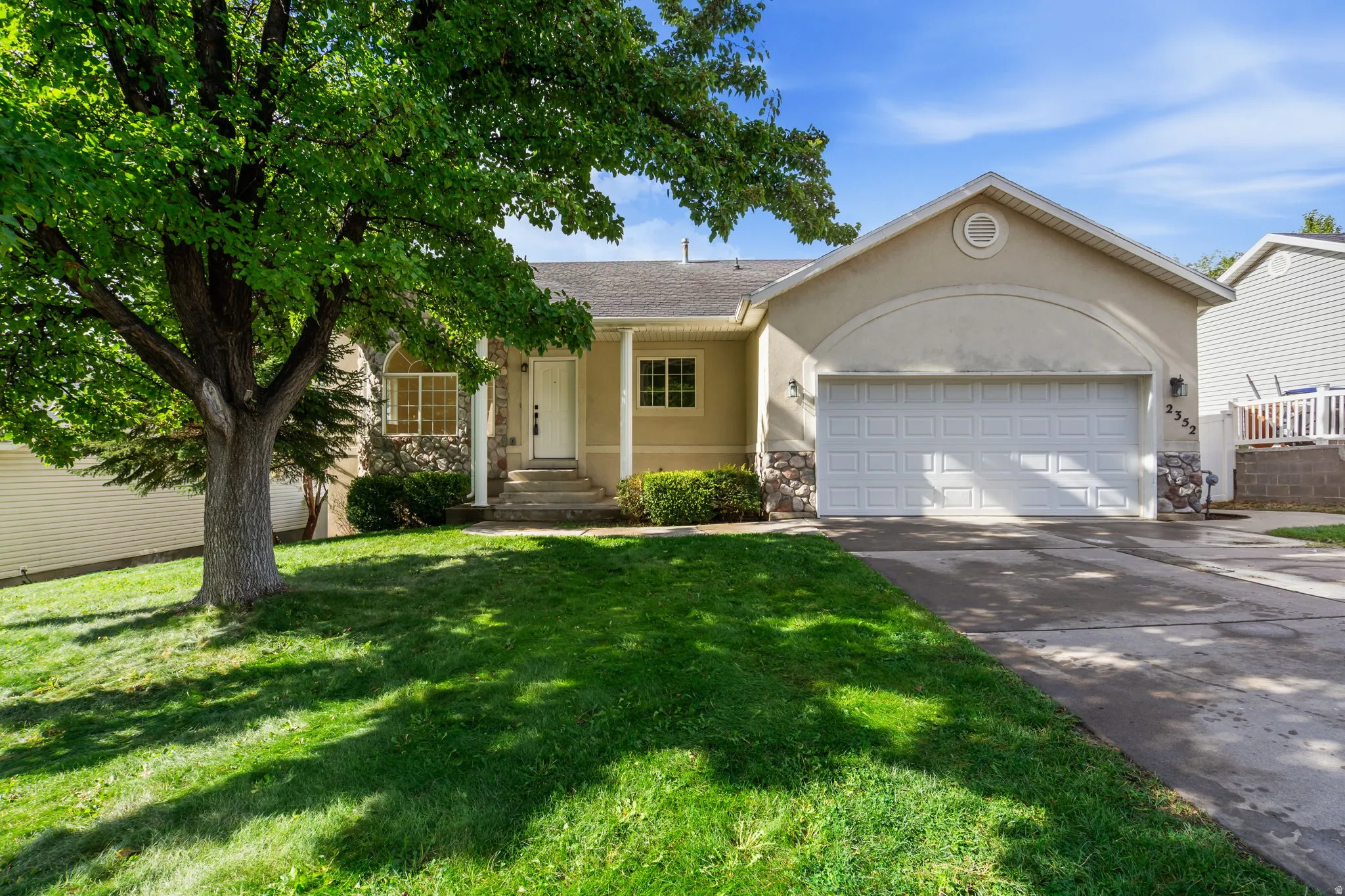 Stucco exterior with stone accents, a prominent two-car garage, mature front yard tree, and an inviting front entrance with covered porch