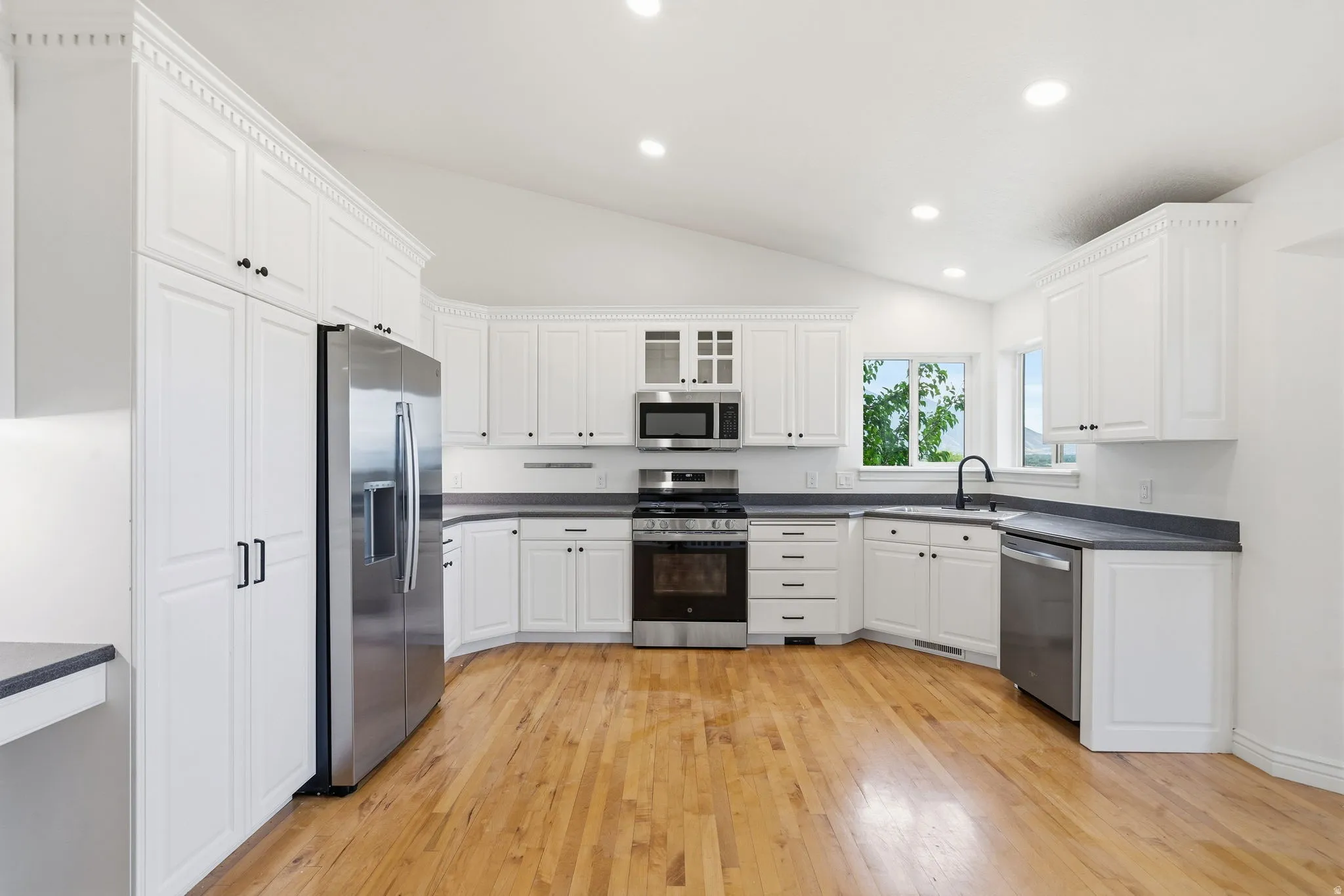 Vaulted ceiling kitchen featuring white cabinetry, wood-finish flooring, stainless steel appliances, dark countertops, and a large window