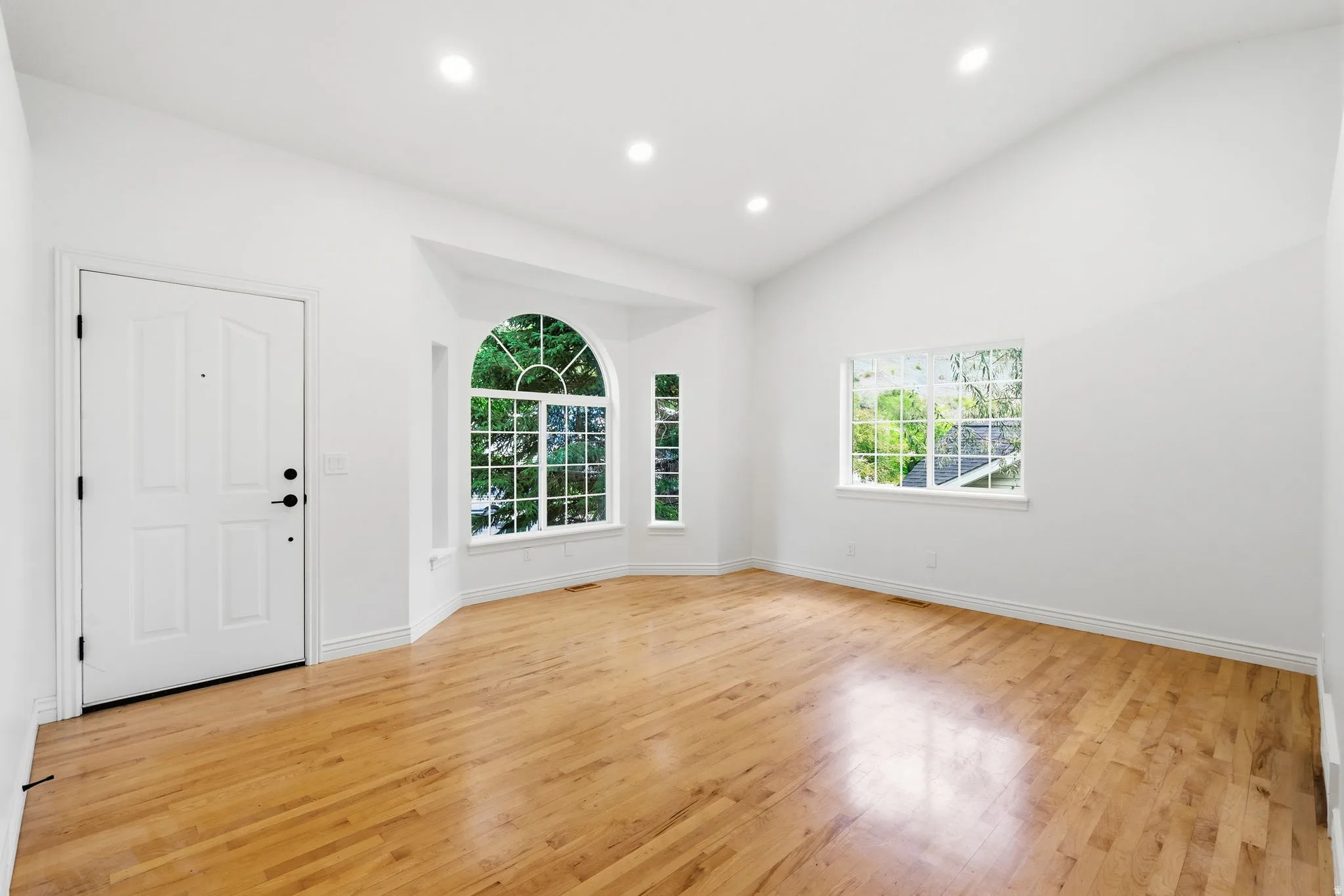 Wood-finish flooring, vaulted ceiling, recessed lighting, white panel door, and a bay window with an arched transom