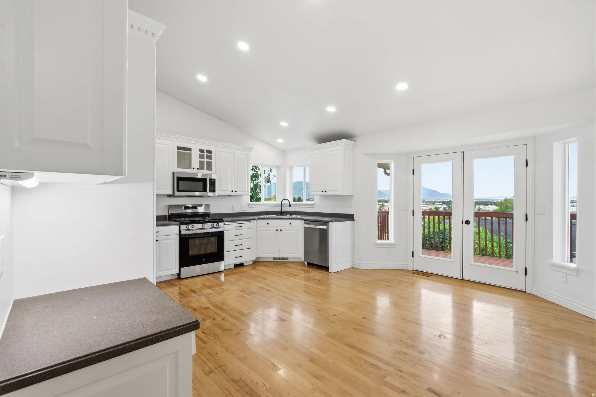 Vaulted ceiling with recessed lighting, light wood-finish flooring, white cabinetry, dark stone-finish countertops, and double glass doors leading to an outdoor deck