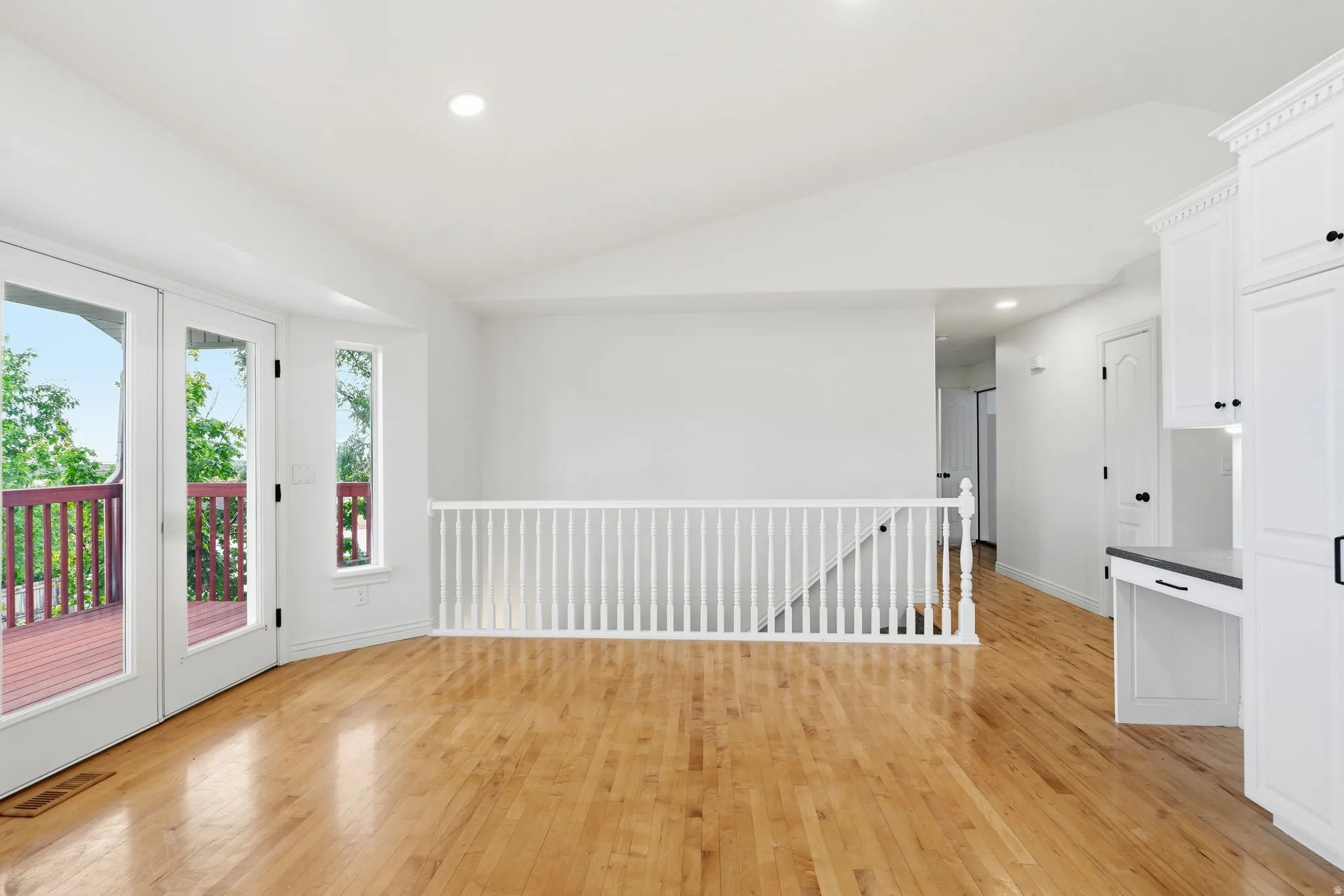 Vaulted ceiling with recessed lighting, light wood-finish flooring, white railing, white built-in cabinetry with black hardware, and glass-paneled French doors opening to an exterior deck