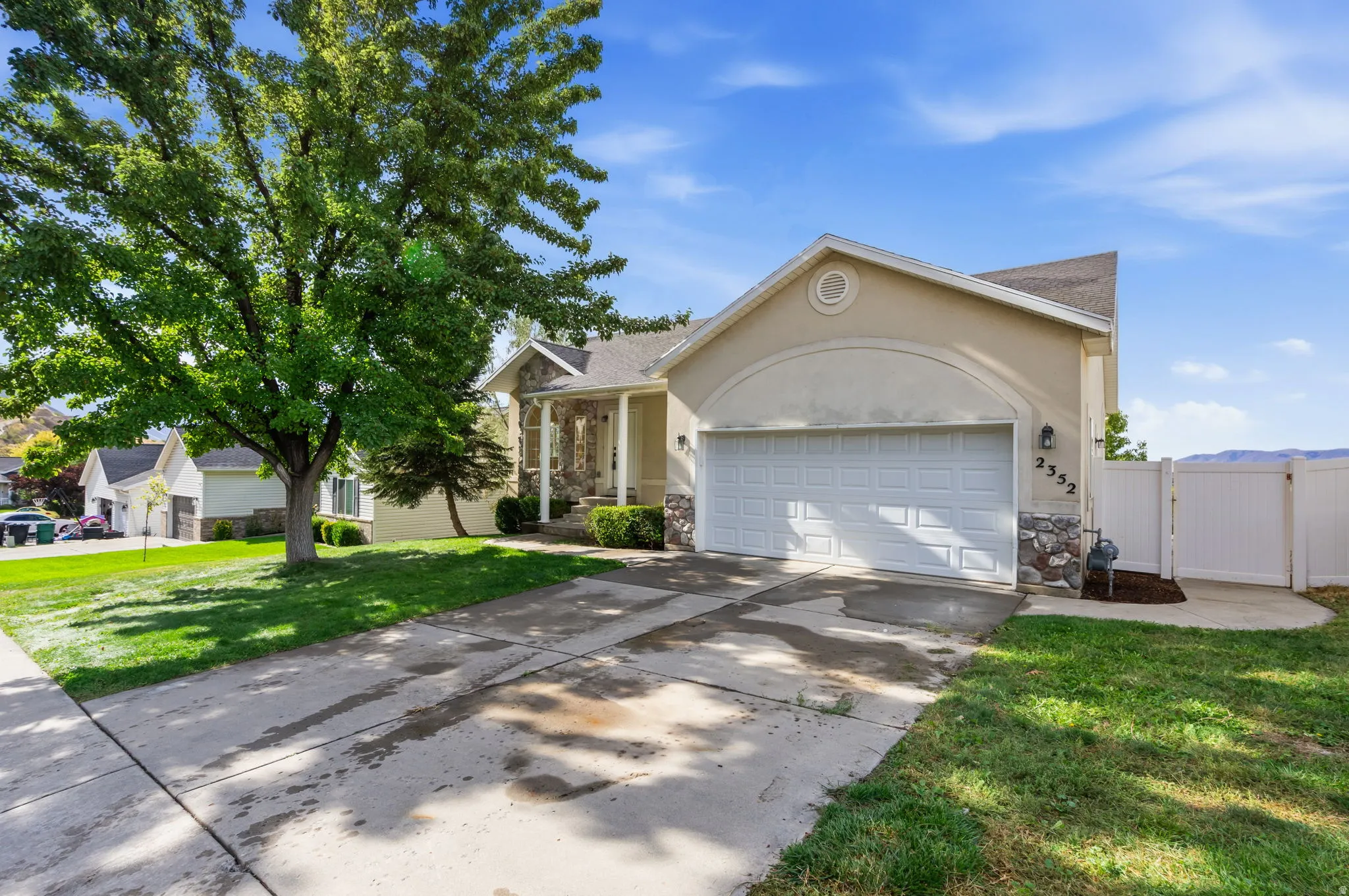 Stucco and stone exterior, attached two-car garage, concrete driveway, front yard with mature tree, and white privacy fencing