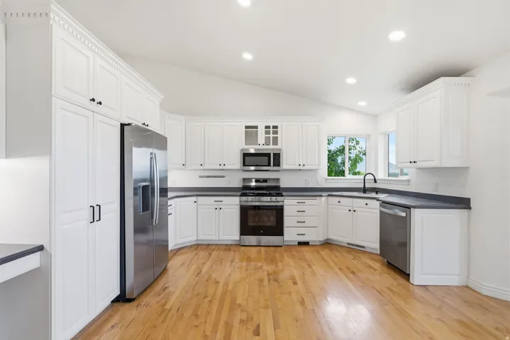 Vaulted ceiling kitchen featuring white cabinetry, wood-finish flooring, stainless steel appliances, dark countertops, and a large window