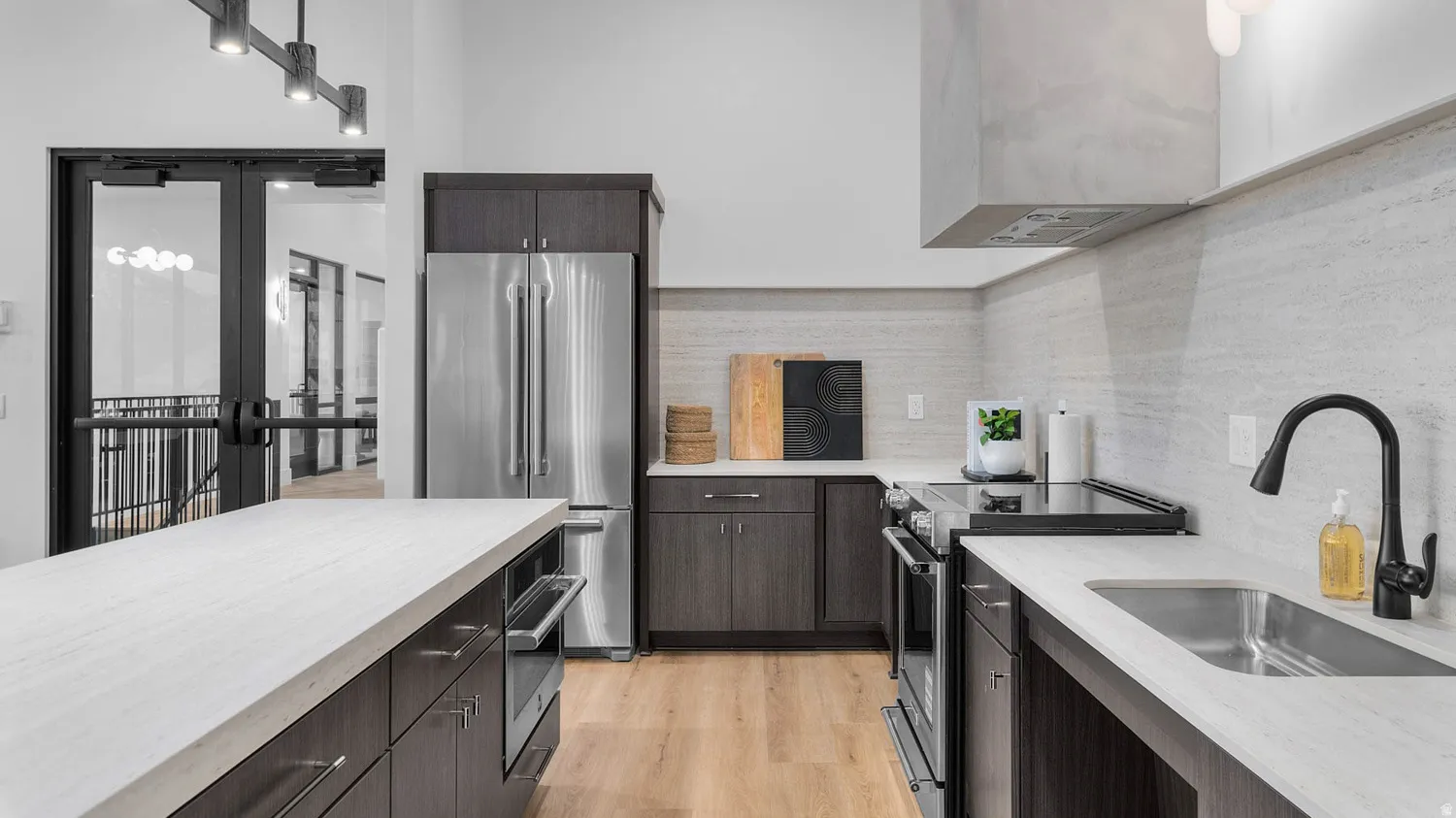 Contemporary kitchen featuring dark wood cabinetry, stainless steel appliances, light-toned countertops, a matching backsplash, and wood-finish flooring