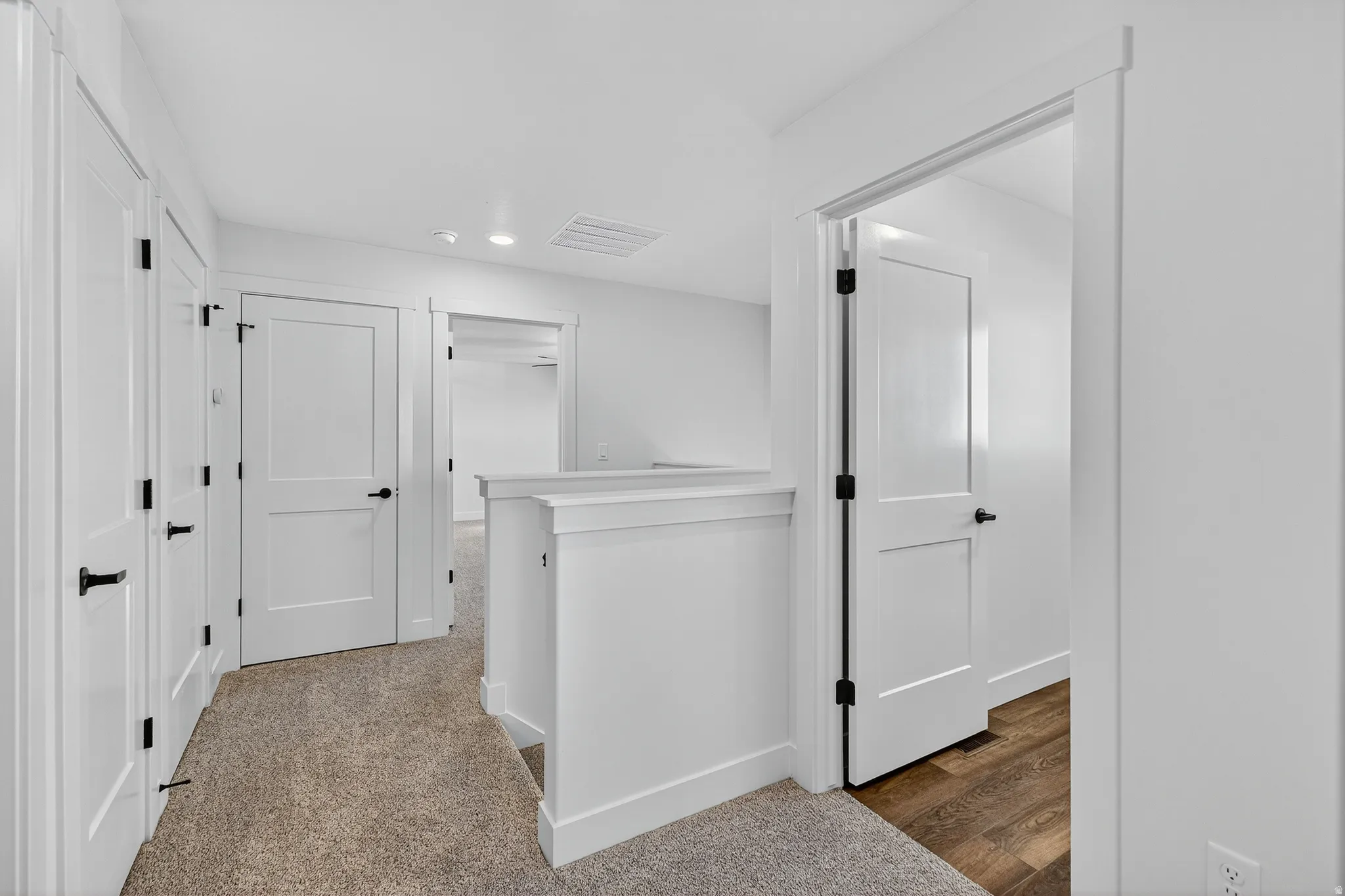 Carpeted hallway with white panel doors and matte black hardware, featuring a half-wall partition and recessed lighting