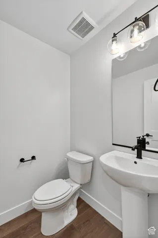 Pedestal sink with matte black faucet, wall-mounted mirror, three-light vanity fixture, white toilet, and wood-finish flooring