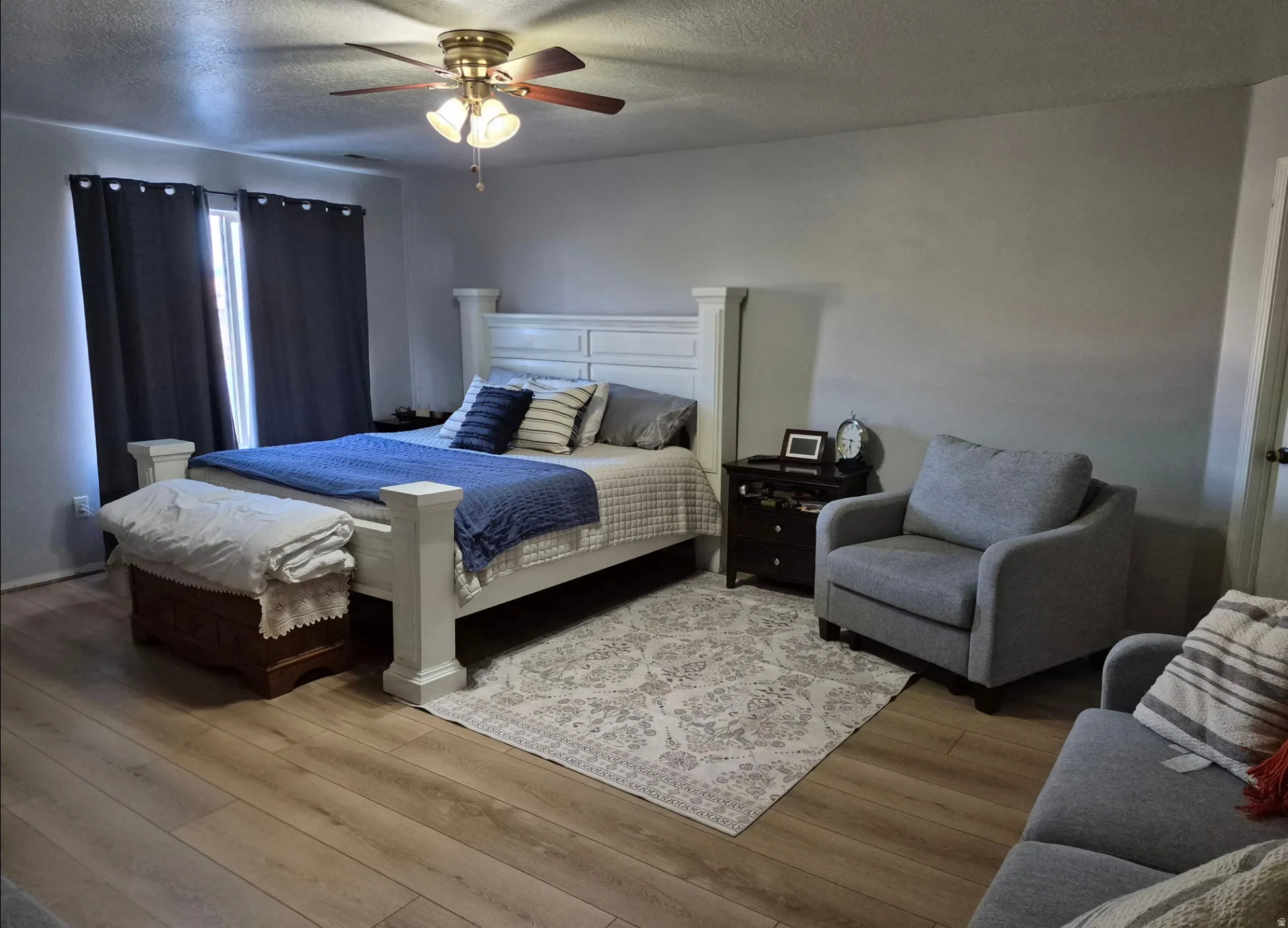 Wood-finish flooring, ceiling fan with integrated lighting, window with dark drapes, light gray wall color, white paneled bed frame