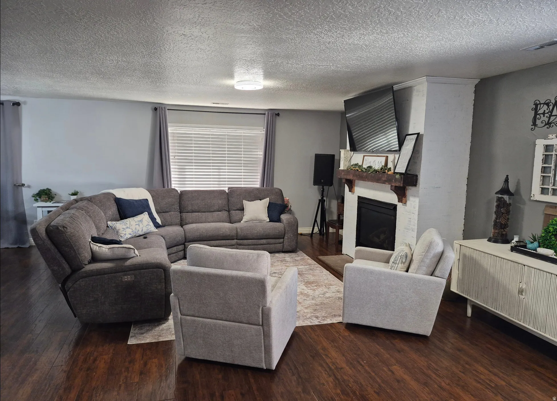 Wood-finish flooring, white brick fireplace with a wood mantel, and a ceiling light fixture