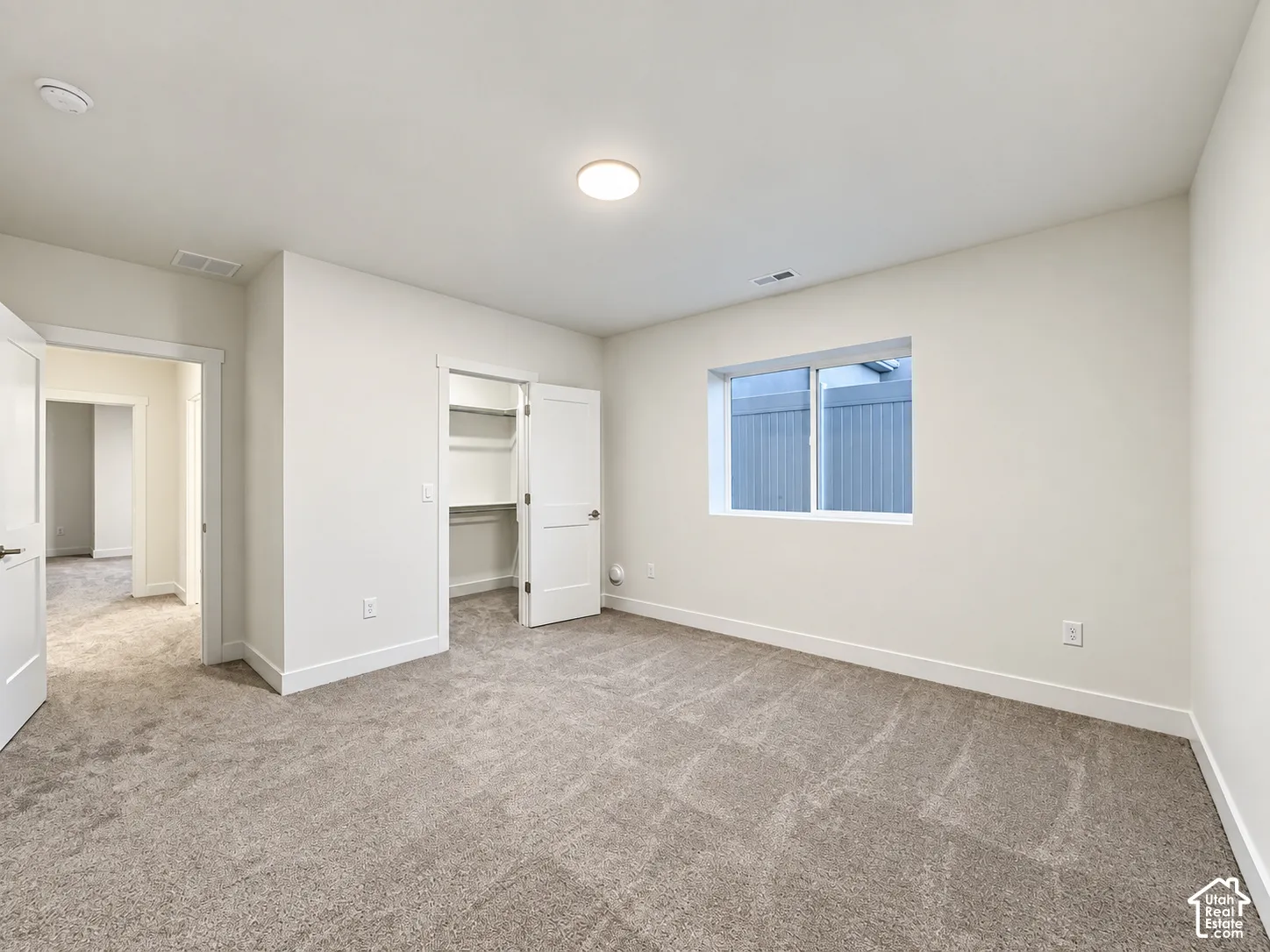Spacious room featuring neutral carpeting, a dual-door closet with shelving, a large window, a ceiling-mounted light fixture, and white trim