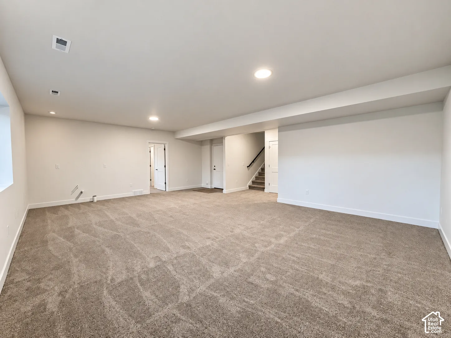 Expansive carpeted area with recessed lighting, white walls, and a stairway featuring dark handrails