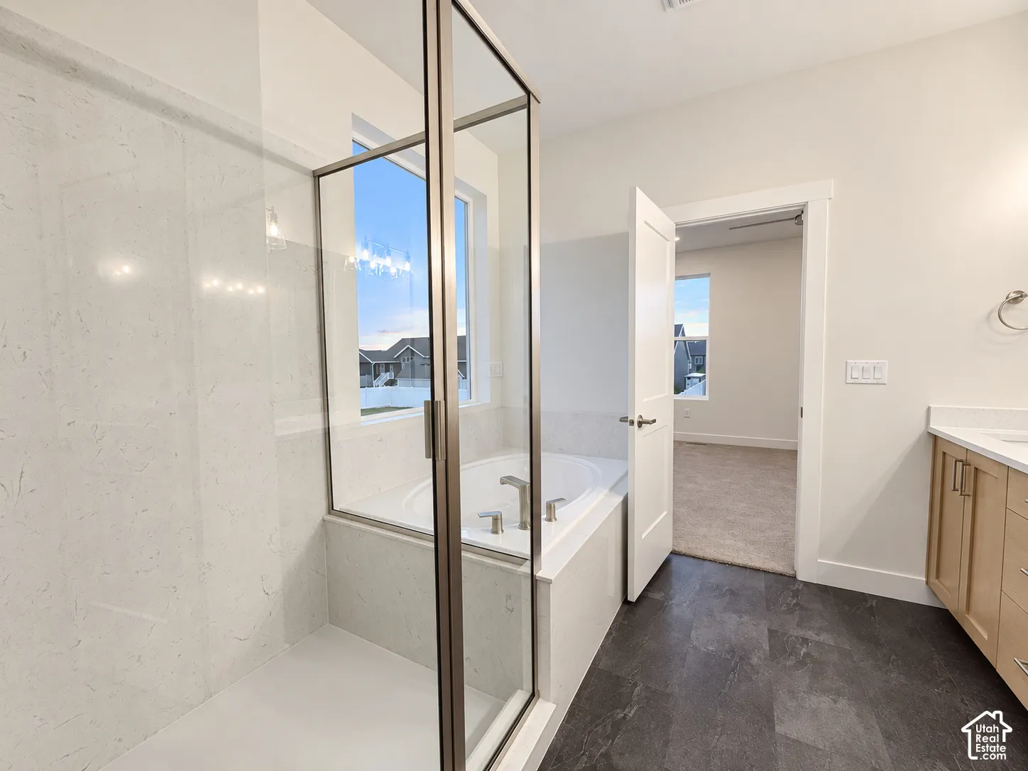 Walk-in shower with clear glass enclosure and tile surround, soaking tub with window, dark-toned flooring, light wood-finish vanity with countertop, and modern fixtures