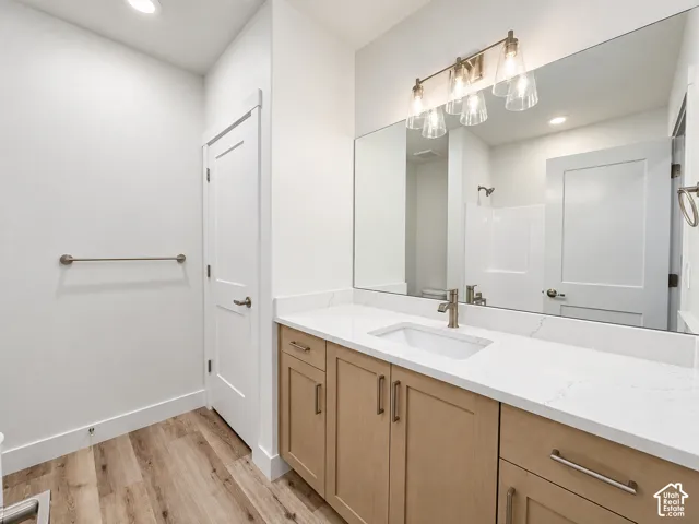 Wood-finish flooring, light wood-tone vanity with brushed nickel hardware, expansive countertop with integrated sink, rectangular wall-mounted mirror, and brushed nickel light fixture with clear glass shades
