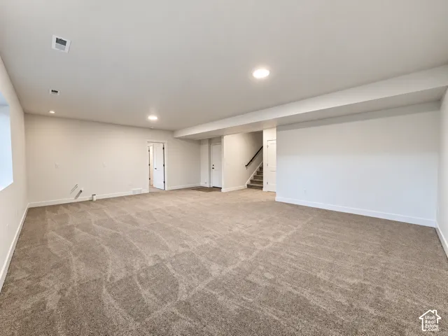Expansive carpeted area with recessed lighting, white walls, and a stairway featuring dark handrails
