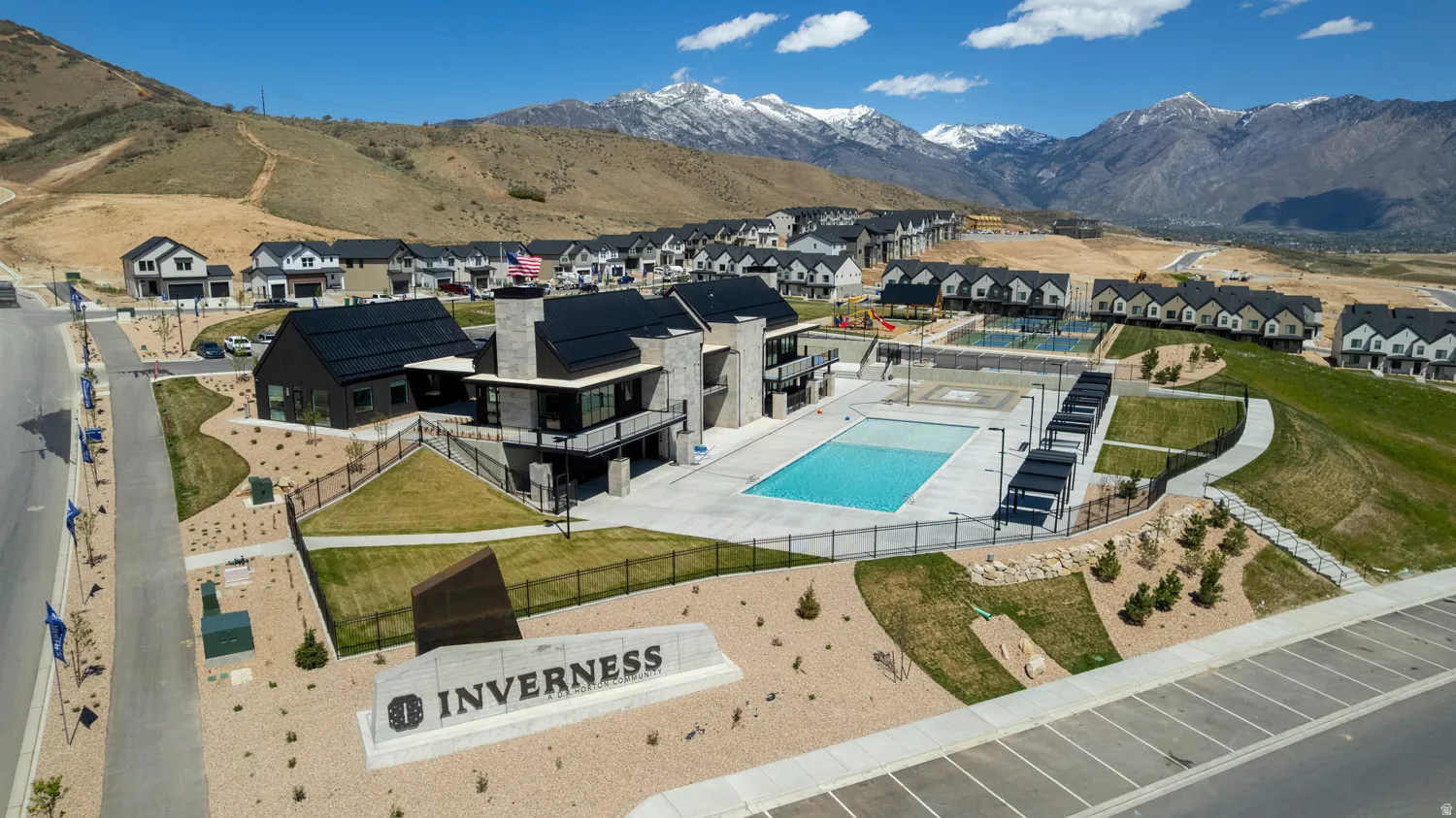 Community entrance with "INVERNESS" monument sign, a modern community building with black roofs, a swimming pool, and a sports court, all surrounded by mountainous terrain