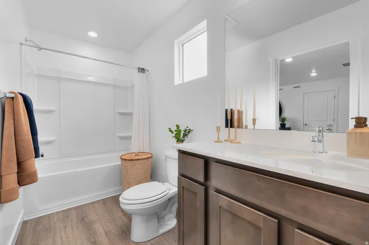 White bathtub and shower combination with integrated shelving, wood-finish flooring, dark wood vanity with integrated sink, light countertop, and wall-mounted mirror