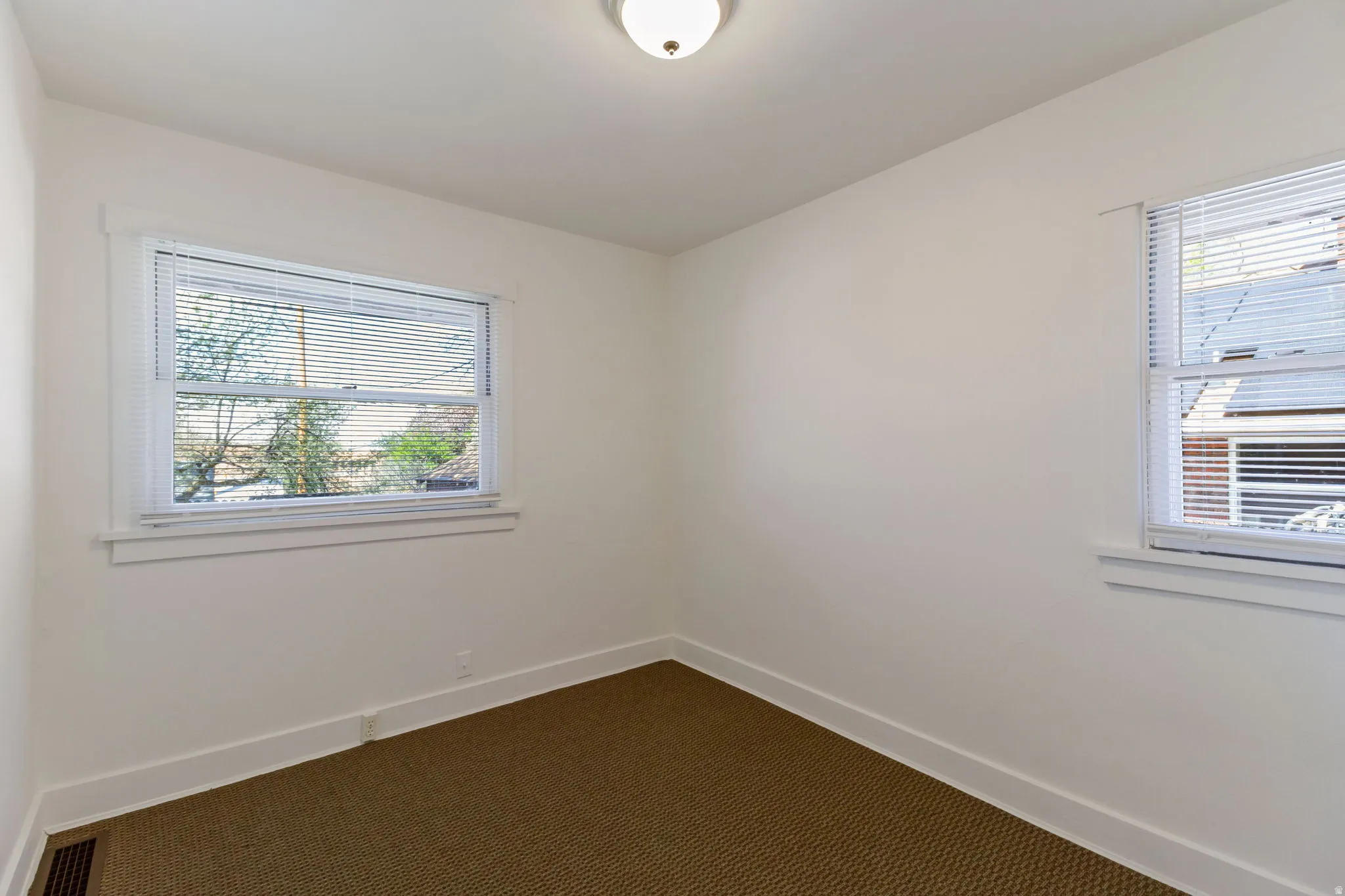White walls, brown textured carpeting, two windows with white blinds, and a flush mount ceiling light fixture