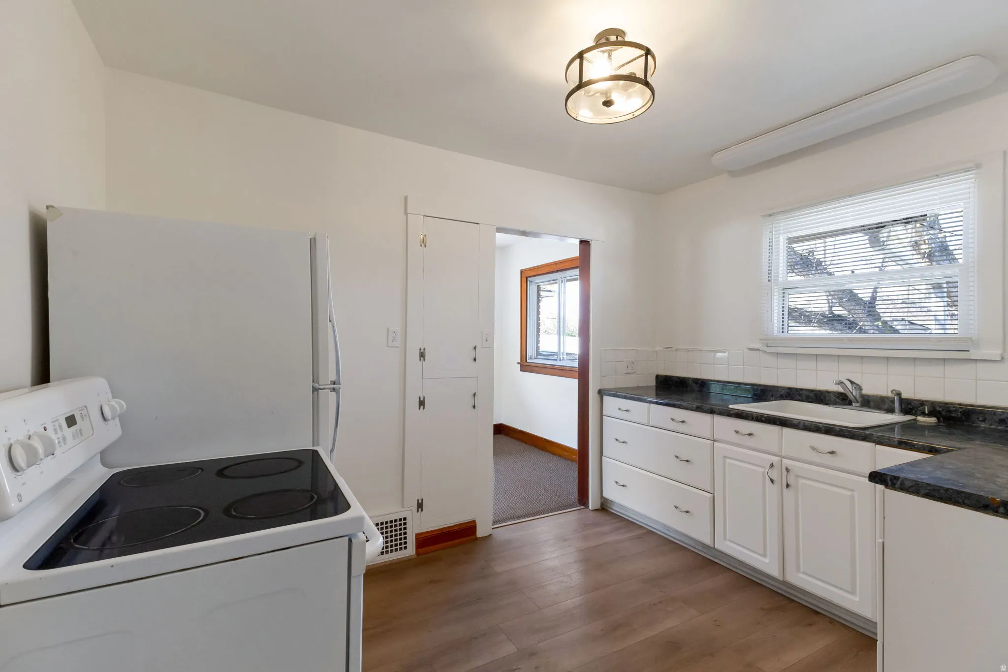 Kitchen featuring wood-finish flooring, white cabinetry, dark countertops, white tile backsplash, and a window above the sink
