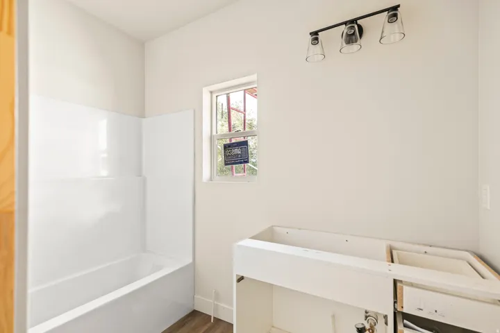 White acrylic tub and shower surround, window with white trim, and a black three-light vanity fixture