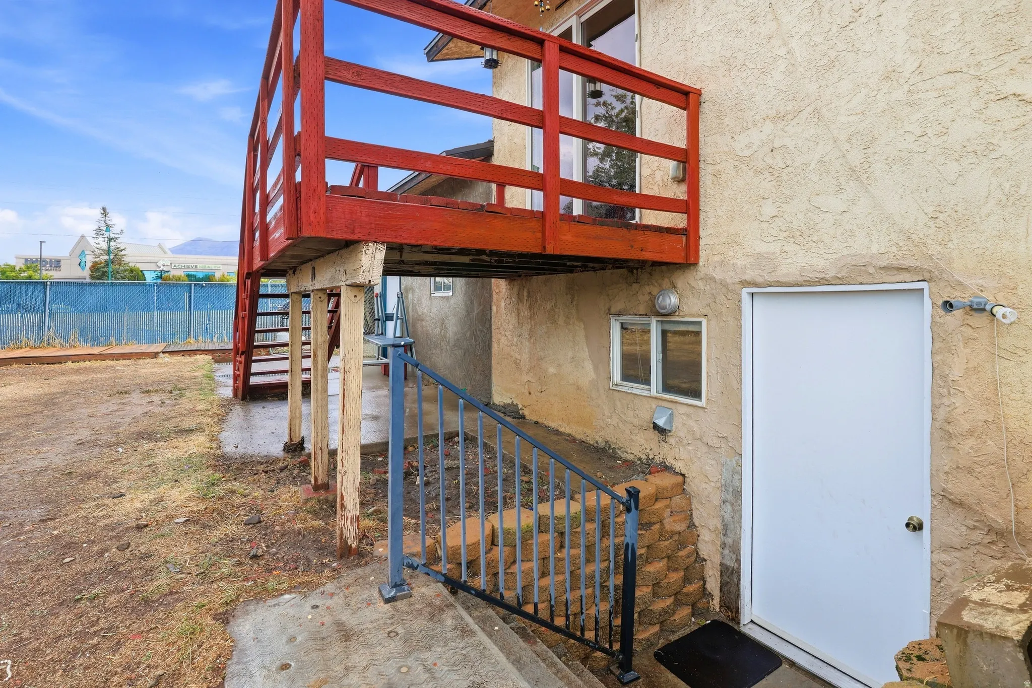 Exterior stucco finish, upper-level wood deck, exterior access door, lower-level window, and a chain-link fence