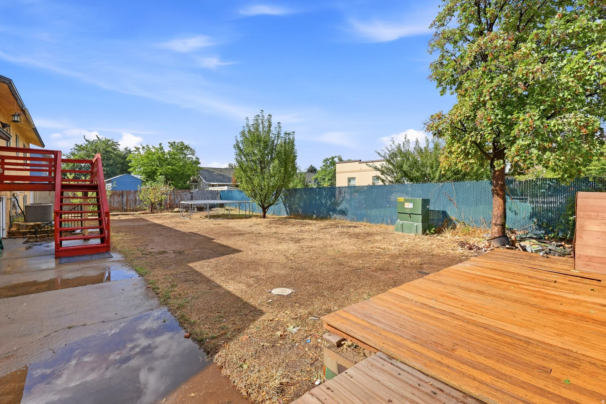 Wood-finish decking with a visible grain texture, expansive backyard space, mature trees, and a ground-level concrete patio