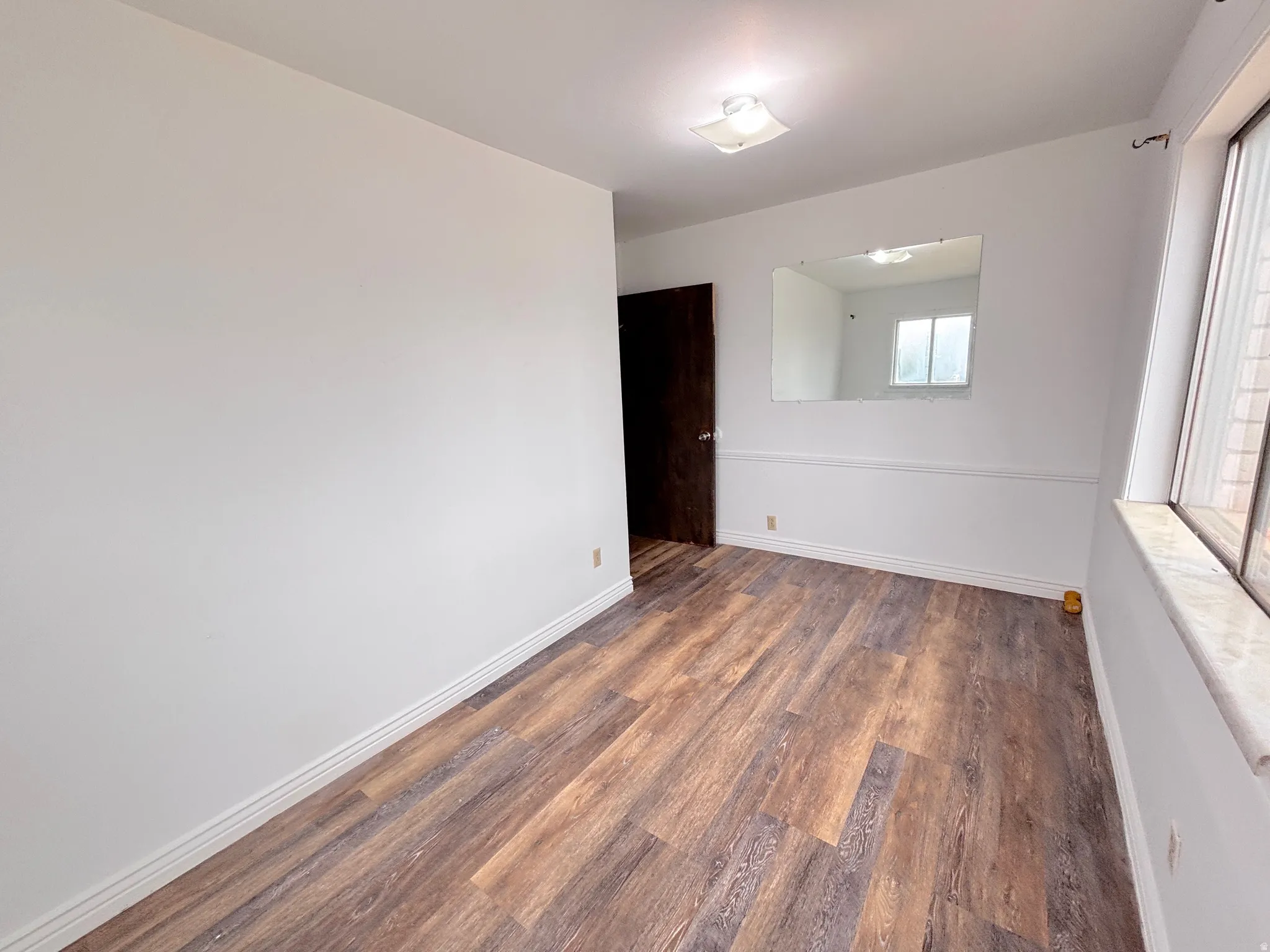 Wood-finish flooring, white walls, dark wood-panel door, square flush-mount light fixture, and a large window with a marble-finish sill