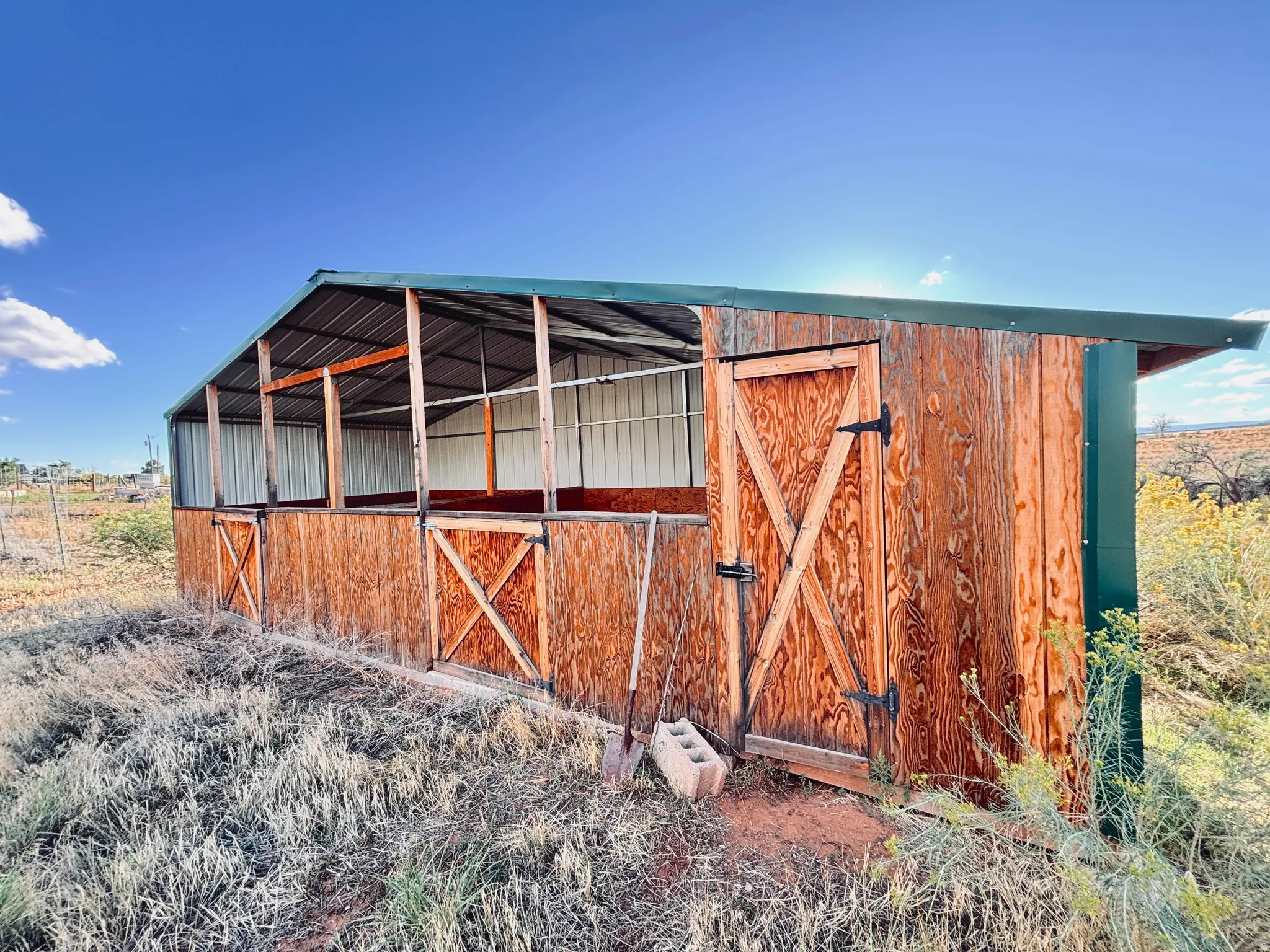 Wood-sided outbuilding with a green metal roof, multiple stalls, and an enclosed storage area