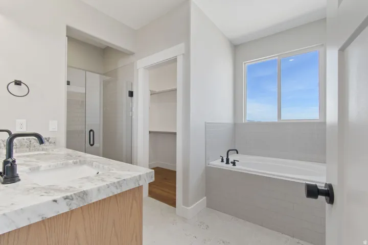 Integrated bathtub with tile surround, walk-in shower with glass enclosure, single vanity with light-toned wood cabinetry, stone countertop, and matte black fixtures, and wood-finish flooring