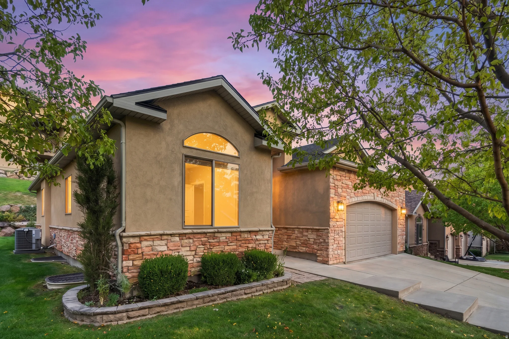 Stucco and stone exterior with an arched garage door, concrete driveway, arched window, and mature landscaping