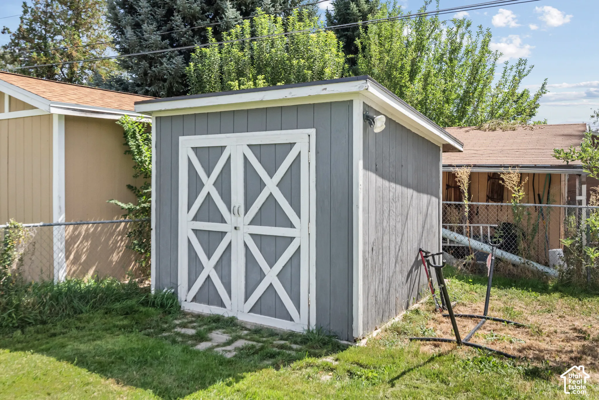 Detached storage shed featuring a gray exterior, white cross-brace doors, and a partial chain-link fence