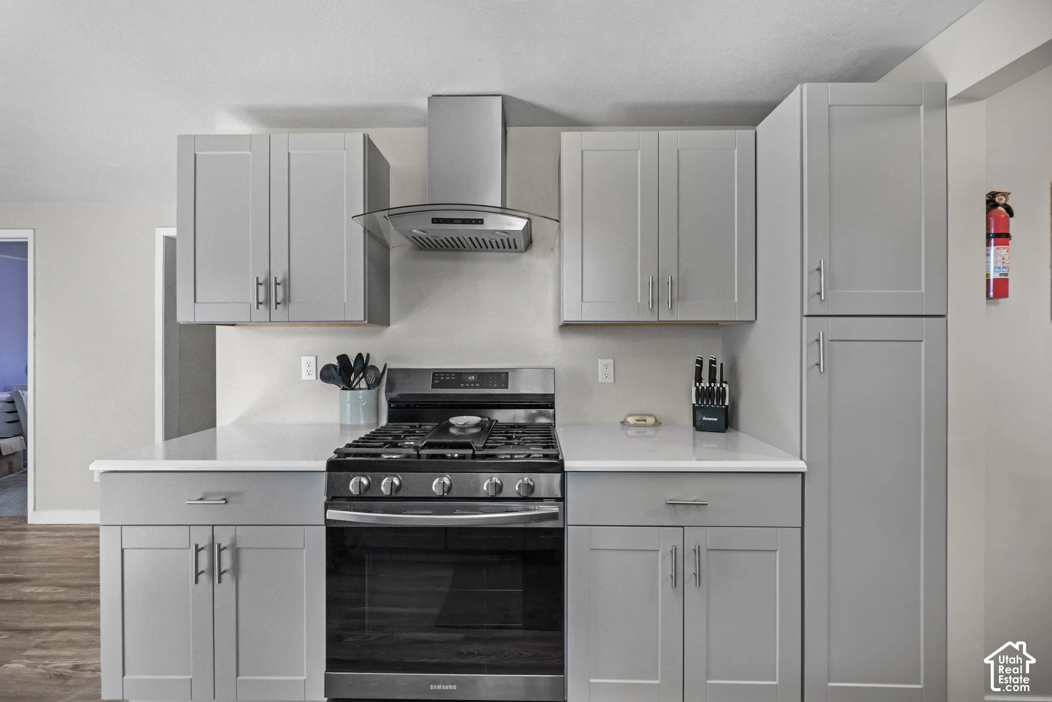 Gray shaker-style cabinetry with brushed nickel hardware, white countertops, stainless steel range, and coordinating range hood