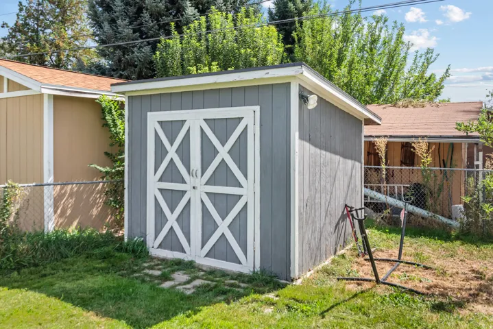 Detached storage shed featuring a gray exterior, white cross-brace doors, and a partial chain-link fence