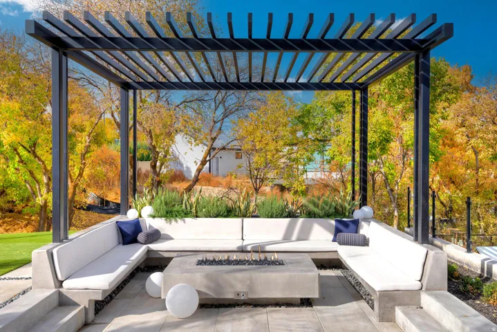 Contemporary pergola with slat roof design, built-in concrete seating, integrated fire pit, and surrounding garden beds