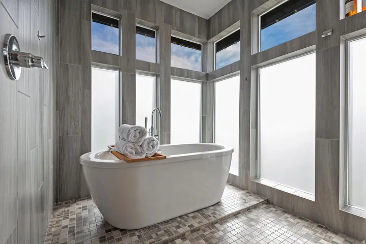 Freestanding soaking tub with chrome fixtures, surrounded by floor-to-ceiling tiling and multiple frosted privacy windows
