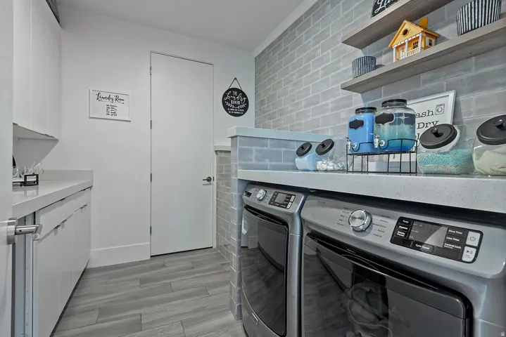 Laundry area featuring wood-finish flooring, white cabinetry, integrated countertop space, and a gray brick-patterned accent wall