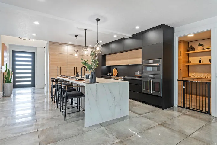 Modern kitchen featuring a large central island with seating, integrated double ovens, warm wood-finish cabinetry, dark flat-panel cabinetry, and a walk-in pantry with shelving