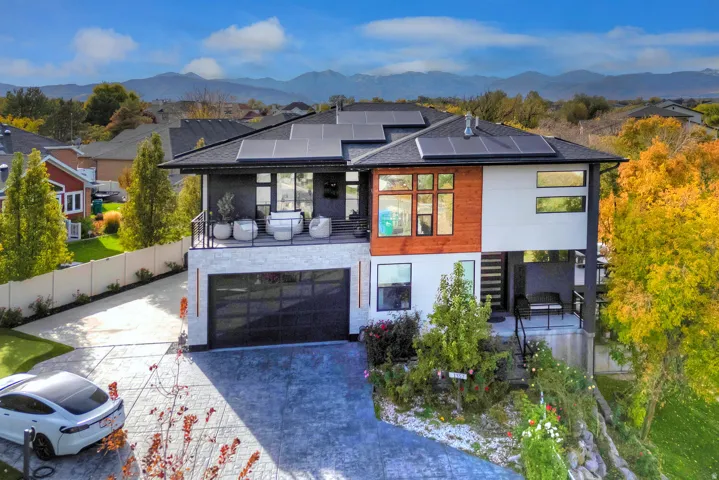 Modern architectural style with a multi-level design, featuring solar panels, a balcony with metal railings, a transparent garage door, and a textured driveway
