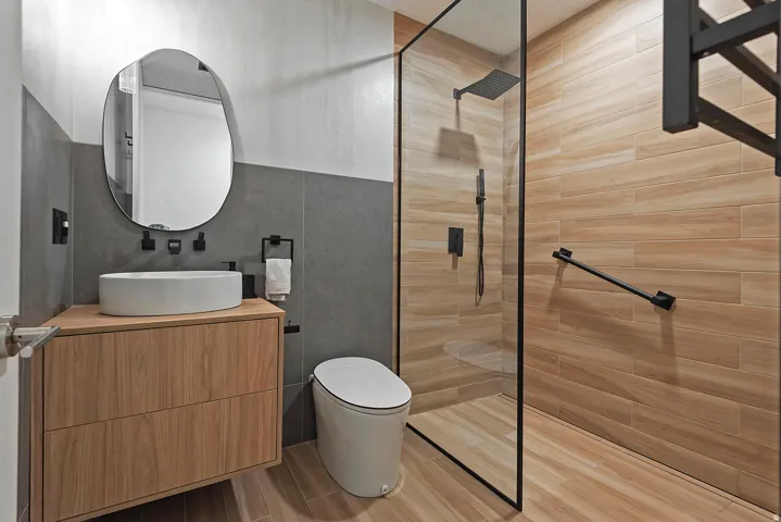 Floating wood-finish vanity with a vessel sink, wall-mounted faucet, and black-framed shower enclosure featuring a rain showerhead