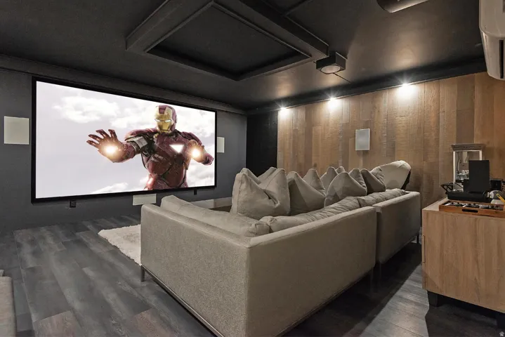 Dedicated media room featuring a large projection screen, wood-finish flooring, accent wall with vertical wood paneling, recessed ceiling lighting, and built-in ceiling speakers