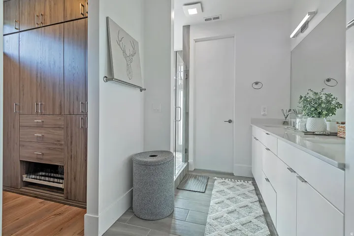 Built-in wood-finish cabinetry, wide plank wood-finish flooring, white vanity with dual sinks, wall-mounted lighting, and a glass-enclosed shower